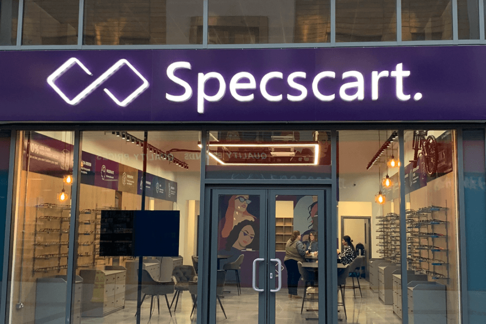 Specscart Urmston store exterior featuring a glowing purple sign, glass entrance, and warm interior lighting with rows of stylish frames on display.