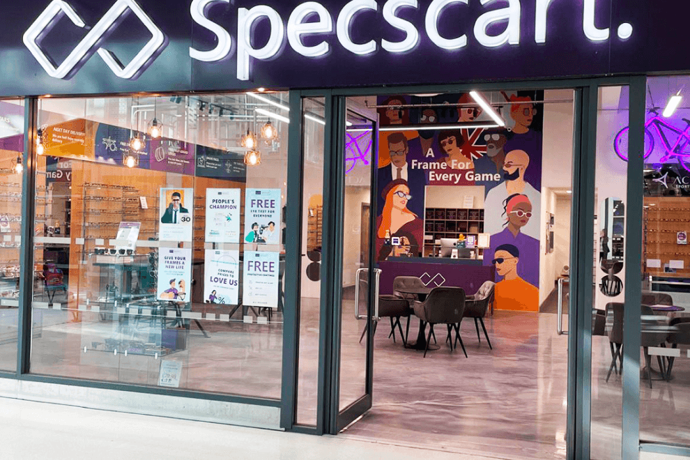Exterior of Specscart Walkden store featuring a glowing logo, sleek glass entrance, and posters advertising free eye tests in the vibrant interior.