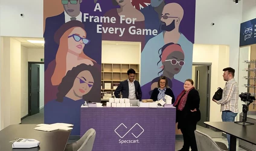Modern Specscart Urmston store interior featuring a large wall mural with the tagline 'A Frame For Every Game' behind a purple reception counter.
