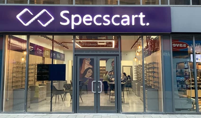 Specscart Urmston storefront at dusk, featuring a large glowing purple sign above glass doors that reveal a warm, modern interior with eyewear displays.