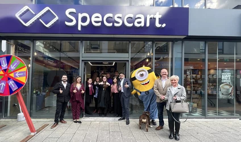 A lively community event at Specscart Urmston featuring staff, a Minion mascot, and a prize wheel outside the modern storefront with a purple sign.