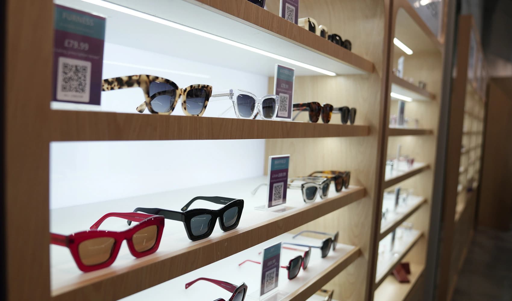 Close-up of stylish sunglasses in tortoiseshell, clear, and red frames displayed on lit wooden shelving with QR code price tags at Specscart Bury.