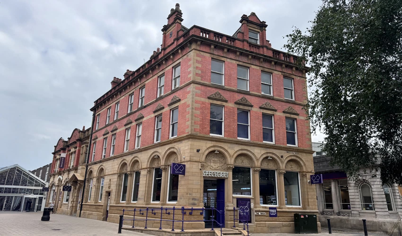 A corner view of the historic Specscart Bury HQ building, showcasing its red brick facade, sandstone arches, and modern purple signage on a clear day.