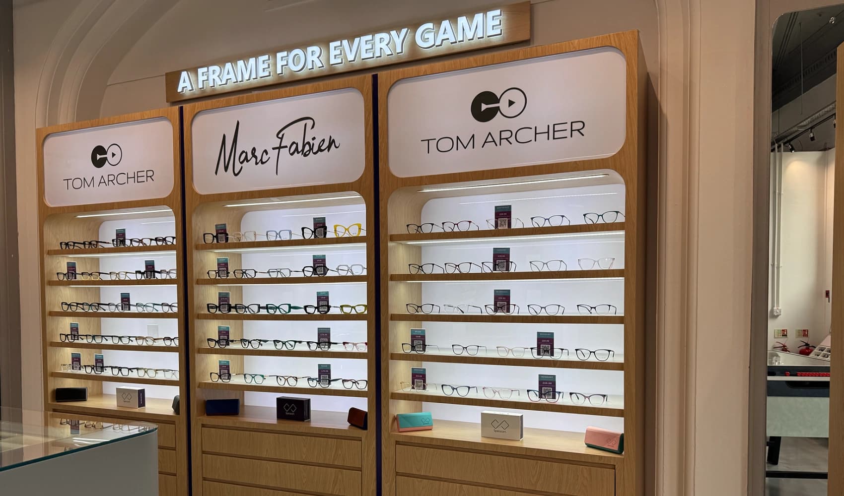 Rows of Tom Archer and Marc Fabien glasses on lit wooden shelves at Specscart Bury, below the illuminated 'A Frame For Every Game' brand tagline.