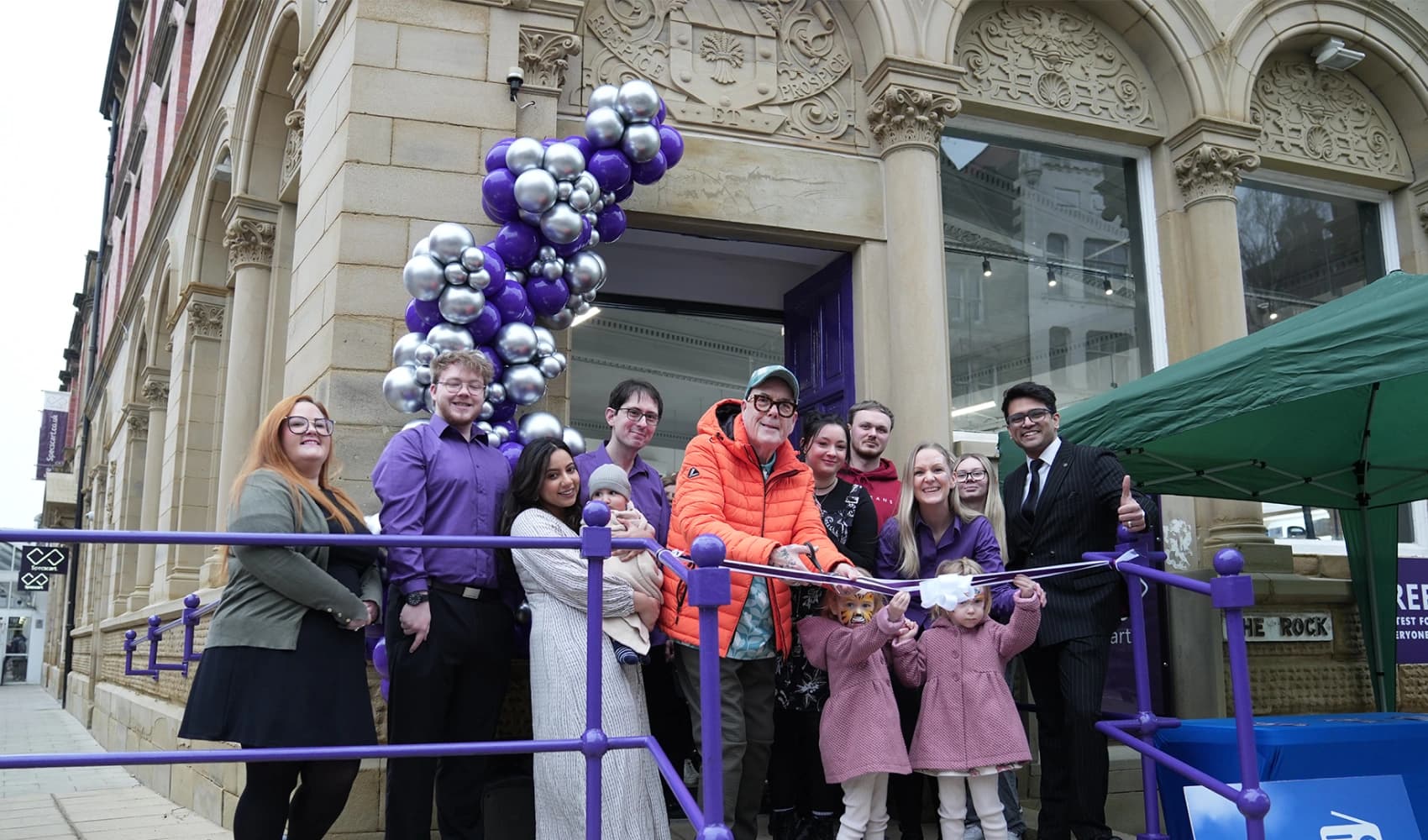 A festive ribbon-cutting ceremony outside Specscart Bury HQ with the Specscart team, decorated with purple and silver balloons.