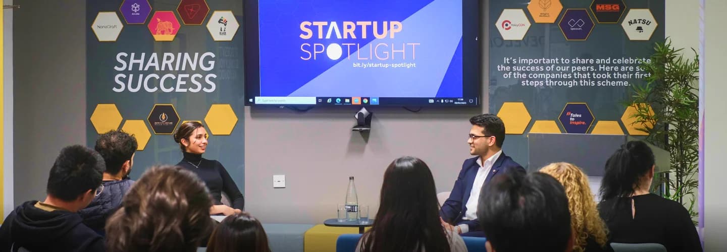 Indoor event with an audience watching two individuals, including a panellist and Specscart founder Sid Sethi in conversation on stage, set against a backdrop featuring the phrases ‘SHARING SUCCESS’ and ‘STARTUP SPOTLIGHT.’ The wall is decorated with honeycomb-shaped panels displaying logos of various companies and motivational messages, highlighting achievement and recognition.