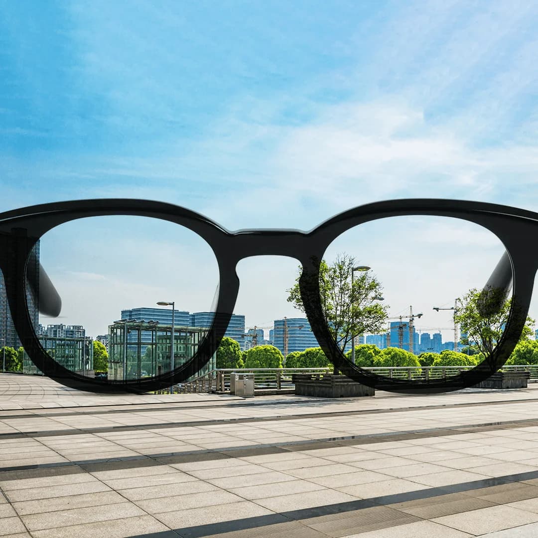 Image of a pair of eyeglass frames placed outdoors with the cityscape in the background. The view seen through the lenses is sharper and clearer compared to the slightly hazy surroundings outside the lens area, illustrating how anti-reflective coatings enhance clarity and minimise distractions for improved vision.