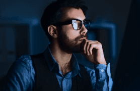 Blue Light Lenses: A man wearing blue light blocking glasses, looking thoughtfully while working on a laptop in a dimly lit setting, representing the benefits of blue light protection.