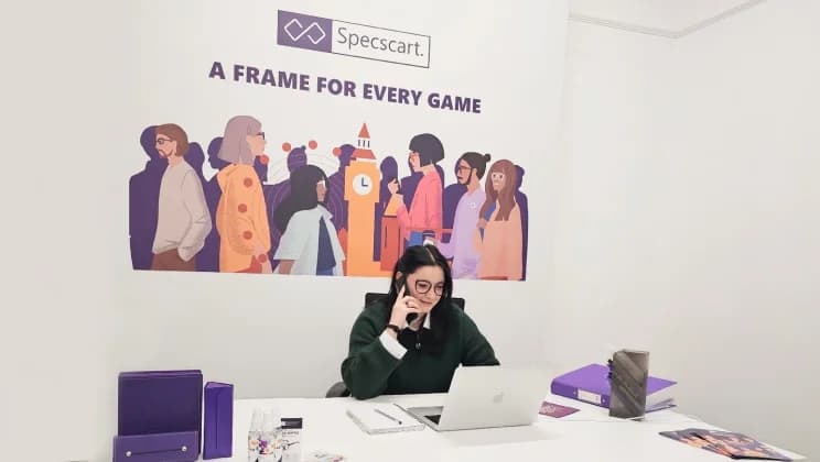 Specscart customer support desk featuring an employee speaking on the phone, seated at a white workspace with a laptop, paperwork, purple folders, and branded accessories. The backdrop displays Specscart’s logo and the slogan ‘A Frame for Every Game’ with a colourful illustration of diverse people.
