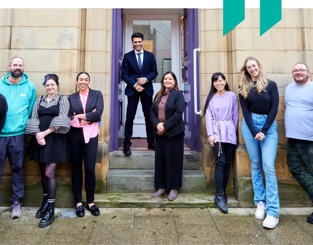 Specscart team members standing together outside the entrance of the company’s Bury headquarters. The group is posed in front of the historic stone building, showcasing a mix of casual and formal attire that reflects an open and friendly workplace culture.