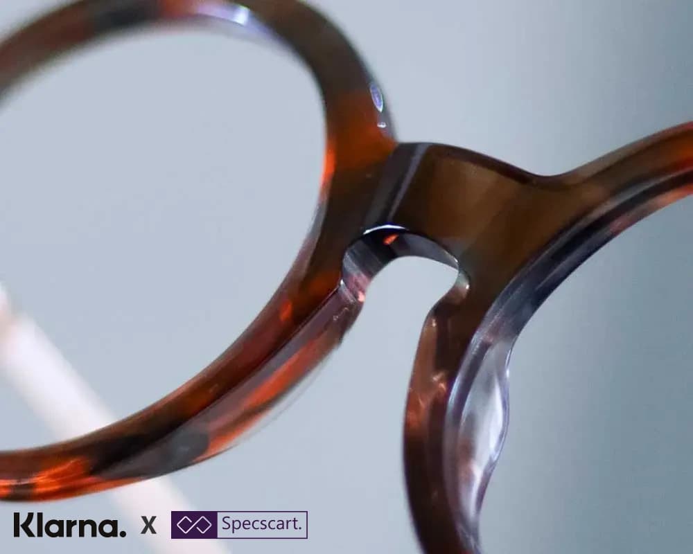 Close-up detail of the bridge and round lenses on a pair of tortoiseshell Specscart glasses. Klarna and Specscart logos are displayed together in the lower left corner, highlighting a payment partnership.