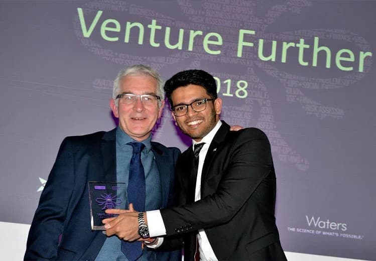 Sid Sethi, Specscart’s founder, in a suit, celebrating the Venture Further 2018 award win on the stage while holding the event trophy. The backdrop features event branding and sponsor details.