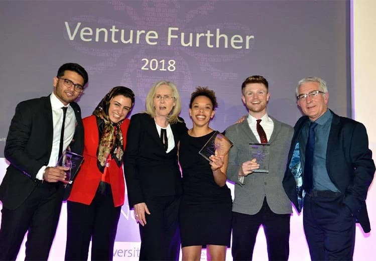 Sid Sethi, Specscart’s founder, standing with a group of attendees on stage at the Venture Further 2018 event. Several participants are dressed in formal attire and holding glass trophies, posing together in front of an illuminated event screen.