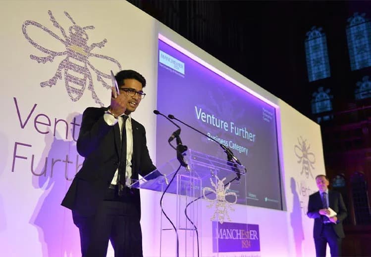Sid Sethi, Specscart’s founder, accepting the Venture Further Business Category Award on stage, raising the trophy at a podium with event branding, Manchester bee symbols, and illuminated signage in a historic venue.