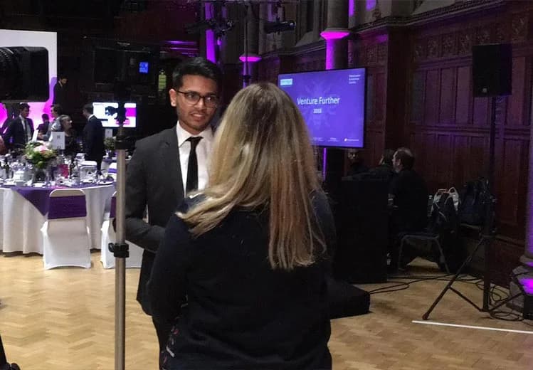Sid Sethi, Specscart’s founder, is being interviewed at the Venture Further 2018 event in a grand hall, with round tables, event lighting, and a stage bearing the official event branding in the background.