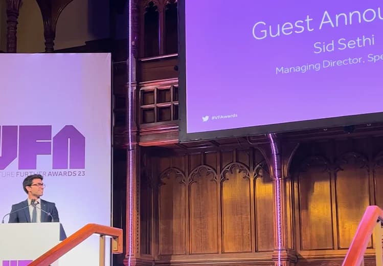 Sid Sethi, Specscart’s founder, standing at the podium to address the audience at Venture Further Awards 2023. The event is set in a grand hall with wood panelling, featuring VFA branding and a projected screen introducing Sid as guest announcer.