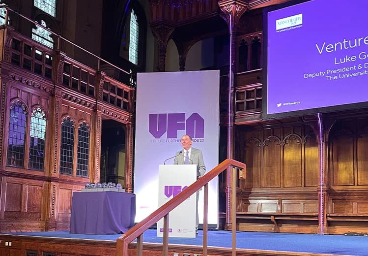 Luke Georghiou, Deputy President & Deputy Vice-Chancellor of The University of Manchester, speaking at the Venture Further Awards 2023 podium. The hall is decorated with VFA banners, a table with trophies, and historic architectural features.
