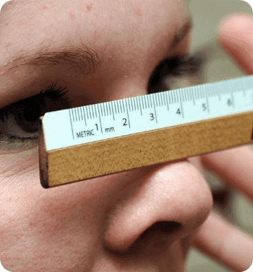 Close-up of a person's eye and nose with a small wooden ruler held horizontally across the bridge of the nose, showing measurement markings for pupillary distance.