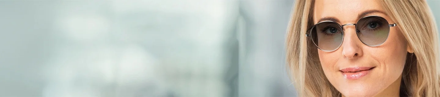 Varifocal Sunglasses A smiling blonde woman wearing elegant round metal sunglasses with gradient varifocal lenses, set against a soft, out-of-focus professional backdrop.