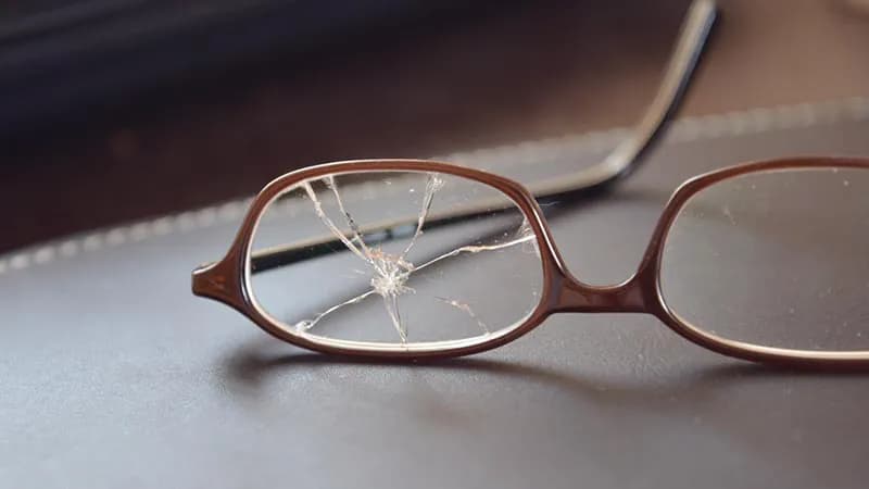 Ultimate Glasses Repair Guide How to fix your broken glasses? Close-up of brown-rimmed glasses resting on a dark surface, featuring a heavily cracked and shattered left lens with a central impact point.