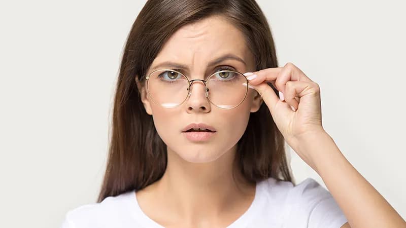 How To Stop Glasses From Sliding Down Your Nose? A woman adjusting her stylish round gold glasses with a puzzled expression, illustrating the common frustration of eyewear sliding down the nose.