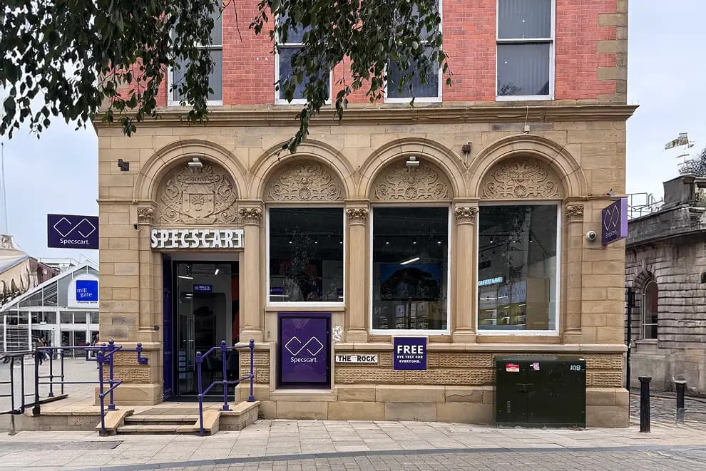 Exterior of Specscart Bury HQ, showcasing a beautiful sandstone facade with ornate arches, purple logo signage, and advertisements for free eye tests.