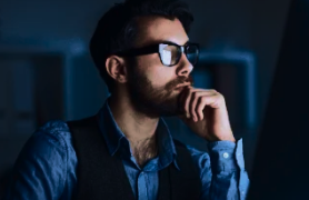 Blue Light Lenses: A man wearing blue light blocking glasses, looking thoughtfully while working on a laptop in a dimly lit setting, representing the benefits of blue light protection.